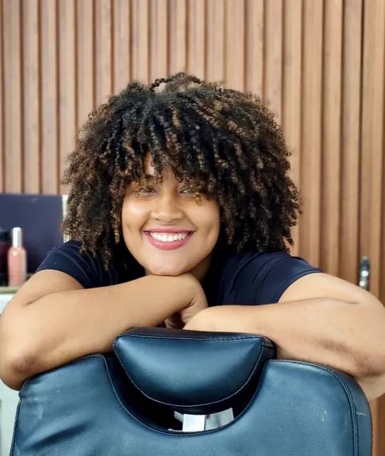 Profissional de beleza sorrindo, apoiada na cadeira do seu salão