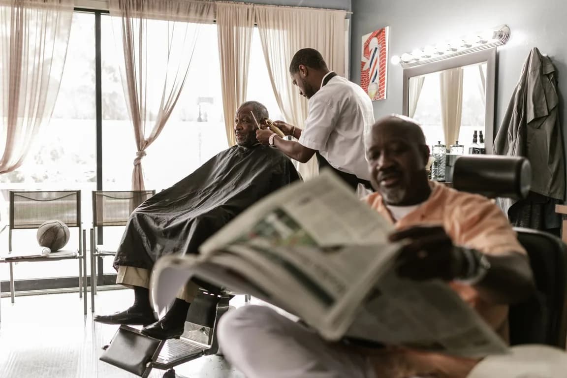 Barbeiro cortando o cabelo de um cliente de capa na cadeira, com outro cliente esperando a vez