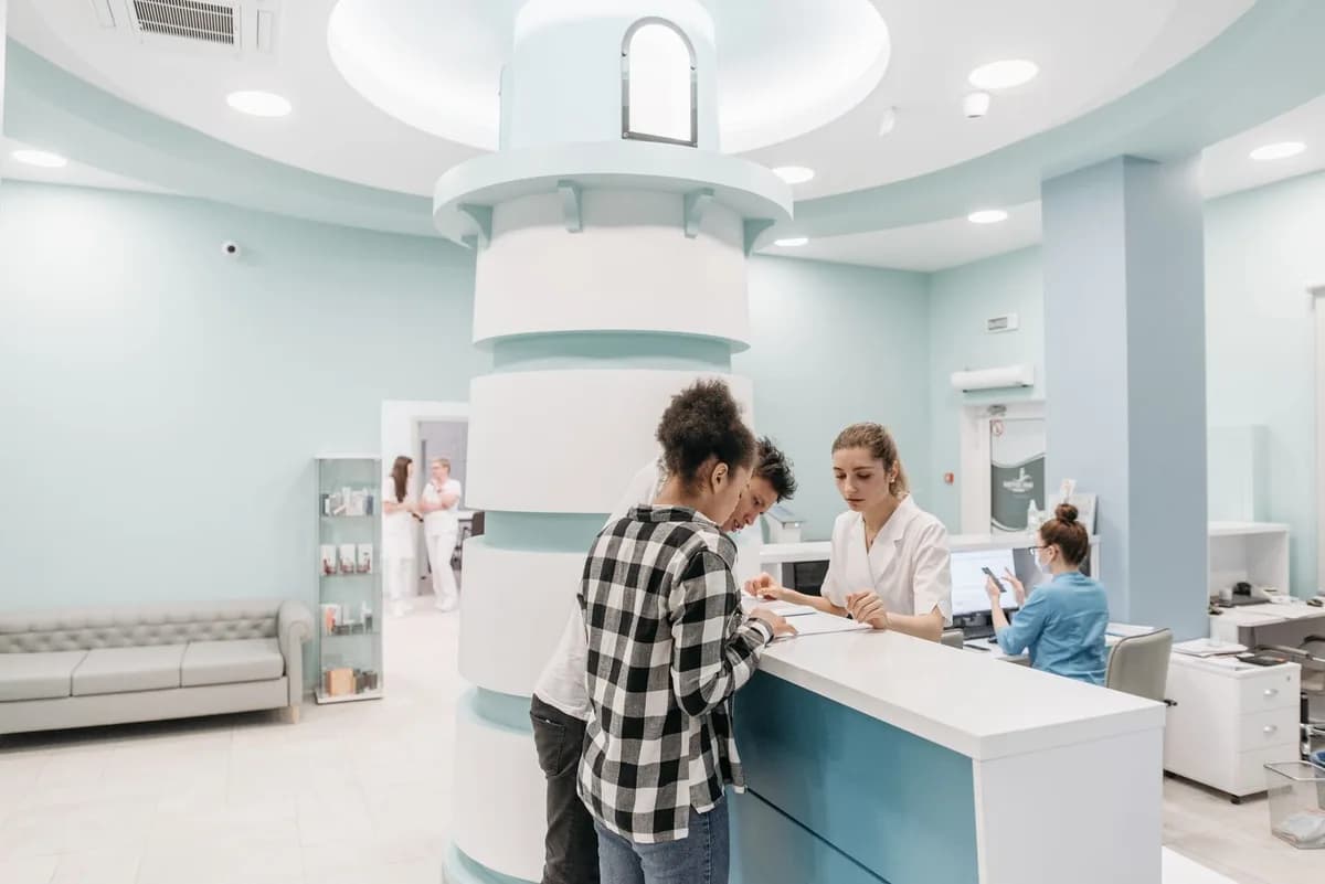 Recepção de clínica com paciente sendo atendida no balcão e profissionais de jaleco ao fundo