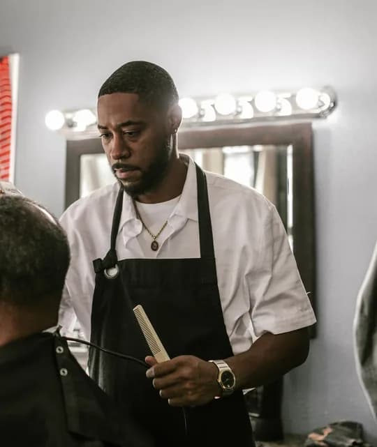 Barbeiro de camisa branca e avental preto, pente na mão, na estação da barbearia